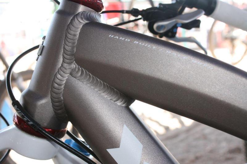 Yeti ASR 5 Alloy: Close-up view of a bicycle frame showcasing the intricate welds on the top tube and head tube, highlighting a metallic gray finish with a matte texture. The image features visible details of the bike's design, including the cable routing and branding.