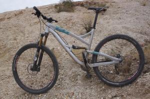 Yeti SB95: A silver and teal mountain bike positioned on a rocky, uneven surface, surrounded by sparse vegetation. The bike features thick, knobby tires suitable for off-road terrain and a sturdy frame designed for rugged trails.