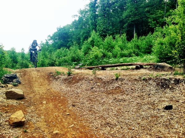 A person riding a mountain bike over a dirt jump in a forested area, surrounded by green trees and small plants. The terrain is uneven with rocks and a wooden ramp visible. Rocky Knob Park mountain bike trail.