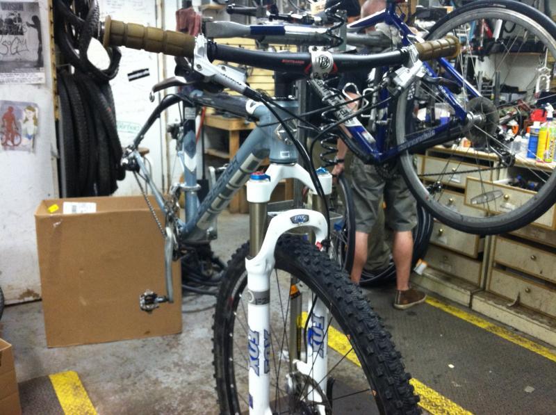 Gary Fisher Paragon: A close-up view of a bicycle in a workshop, featuring a prominent front fork with a suspension system and handlebars. In the background, another bicycle is partially visible, along with various tools and equipment. The space appears cluttered, typical of a bike repair shop.
