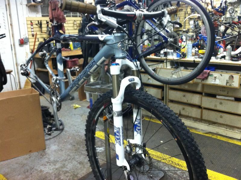 Gary Fisher Paragon: A partially disassembled mountain bike elevated on a work stand in a bike repair shop. The bike features a gray frame, front forks with white suspension components, and knobby tires. Tools and other bicycles are visible in the background, indicating an active workshop environment.