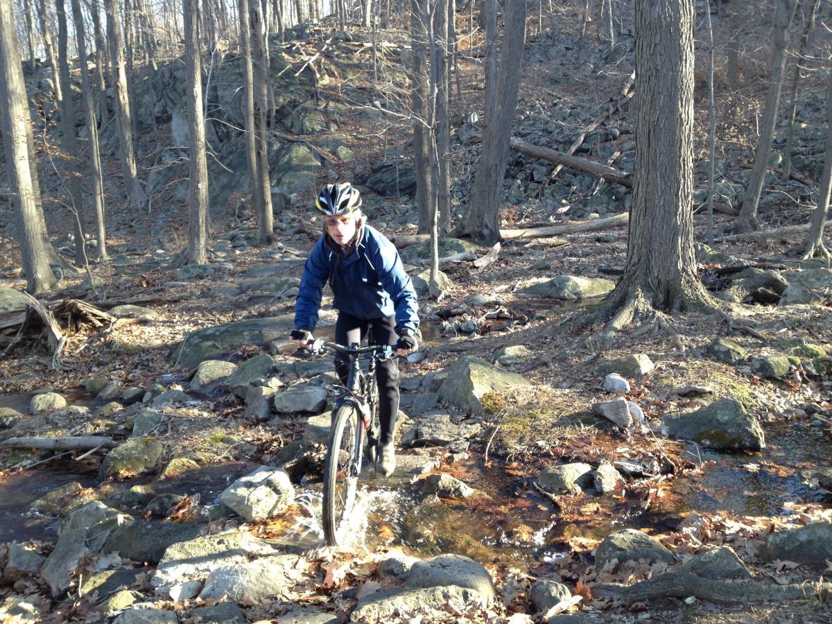 A person riding a mountain bike through a rocky, wooded area, navigating over stones and a small stream. The landscape features tall trees and a rugged terrain, indicating an outdoor trail. The individual is dressed in outdoor gear, including a helmet and a blue jacket, focused on maintaining balance while cycling. Ringwood Skylands Manor mountain bike trail.