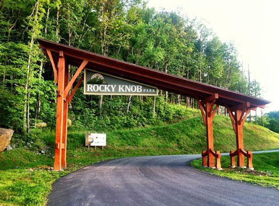 "Entrance sign for Rocky Knob Park, featuring wooden supports and a green landscape in the background with trees. A paved road leads into the park." Rocky Knob Park mountain bike trail.