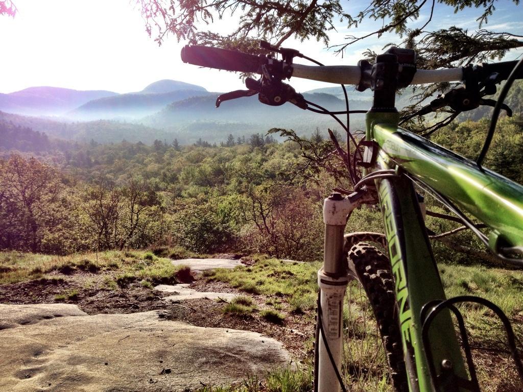 A close-up view of a green mountain bike positioned on a rocky outcrop, overlooking a lush, misty valley and distant mountains under a bright sky. The bike’s handlebars and front fork are in focus, while the background showcases vibrant greenery and soft, rolling hills. Panthertown Valley mountain bike trail.