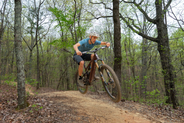 A cyclist in mid-air jumps over a dirt path surrounded by trees in a forested area. The rider wears a helmet and a blue t-shirt, showcasing their skills on a mountain bike. Coldwater Mountain mountain bike trail.