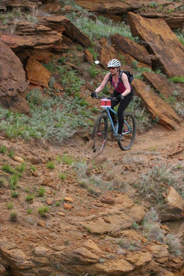 A mountain biker navigating a rocky trail surrounded by earthy, rocky terrain and patches of grass. The cyclist is wearing a helmet and a backpack, and is focused on maintaining balance while biking uphill. Switchgrass mountain bike trail.