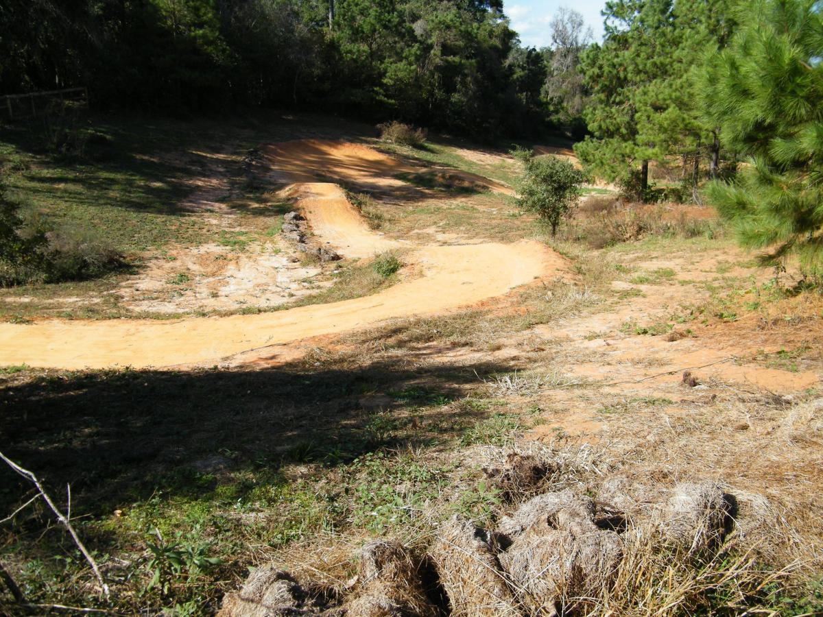 A winding dirt path curves through a grassy area surrounded by trees, with patches of dried grass and scattered shrubs. The terrain is uneven, showcasing a mix of soil and vegetation in a natural setting. Tom Brown / Lafayette Heritage Park mountain bike trail.
