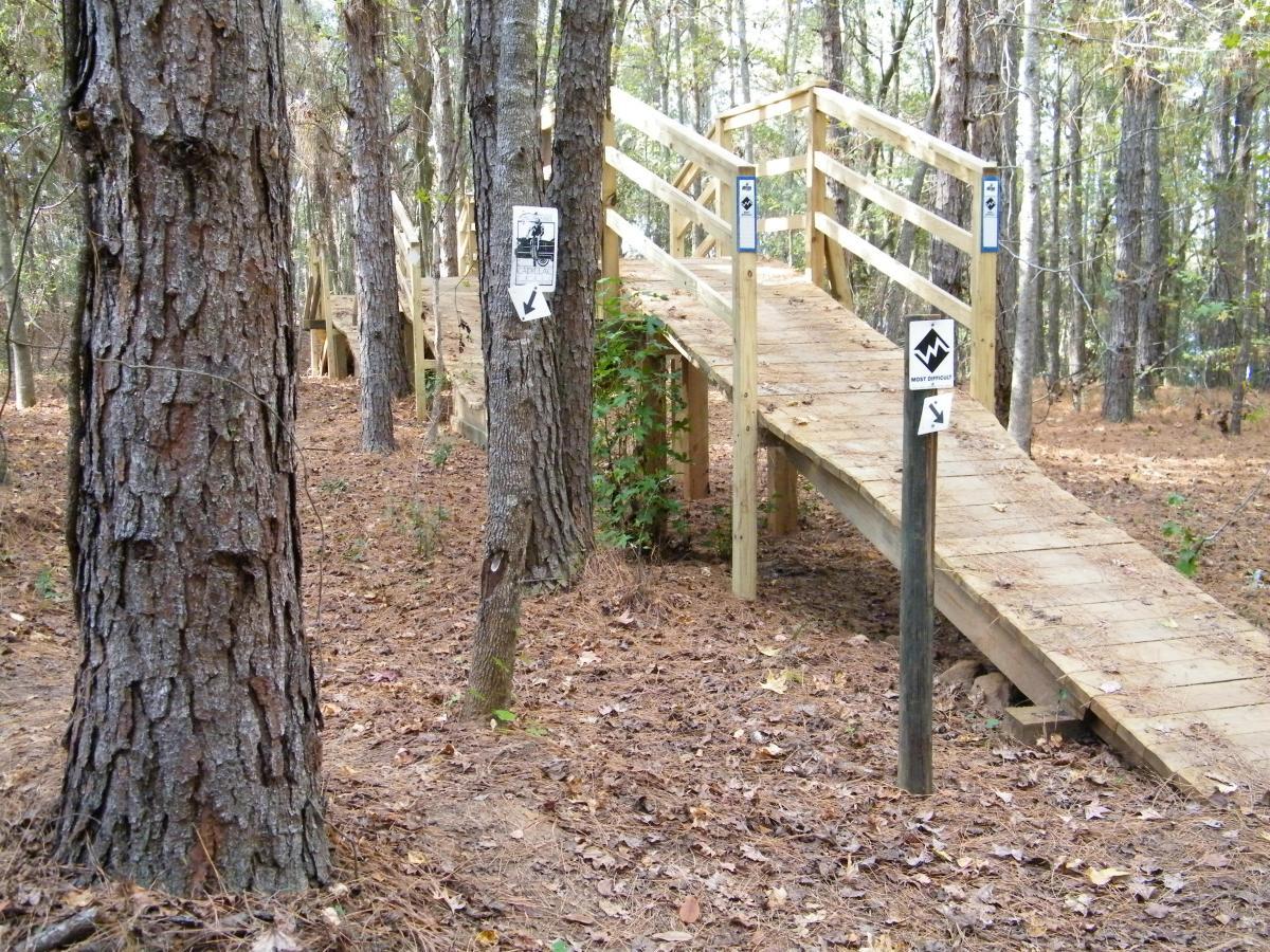 A wooden bridge surrounded by trees in a forested area, with signs indicating trail directions and difficulty levels. The ground is covered with pine needles and fallen leaves. The bridge has handrails and leads into the woods, suggesting a hiking or nature trail. Tom Brown / Lafayette Heritage Park mountain bike trail.