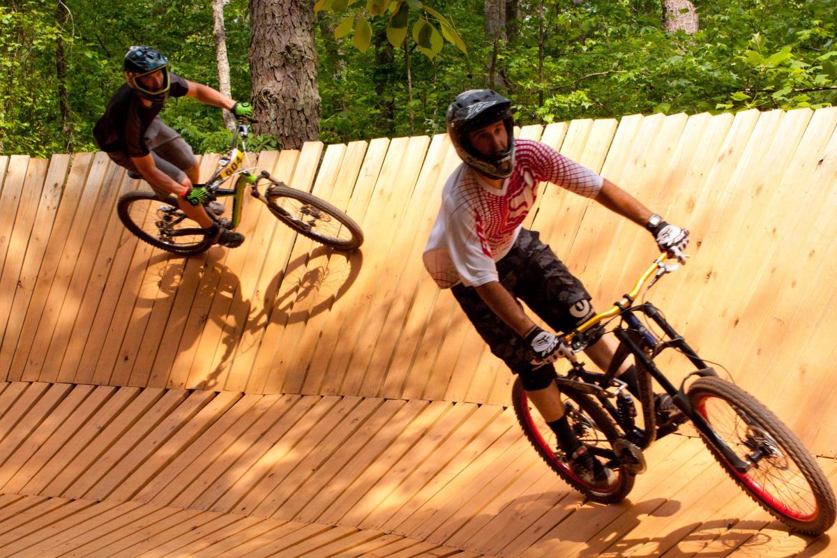 Two mountain bikers maneuvering on a wooden pump track surrounded by trees. One rider is wearing a black helmet and green gloves, leaning into a turn, while the other, in a white and red jersey, follows closely behind. The track features a series of wooden slats, undulating in a wave-like pattern. Quehl Holler mountain bike trail.