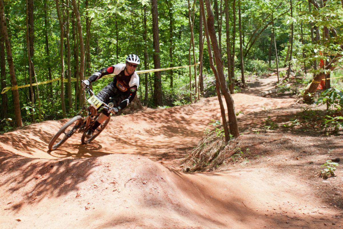 A mountain biker racing on a dirt trail in a forested area, leaning into a sharp turn. The rider is wearing a helmet and protective gear, with a race number displayed on their bike. Sunlight filters through the trees, illuminating the dirt path and surrounding greenery. Quehl Holler mountain bike trail.