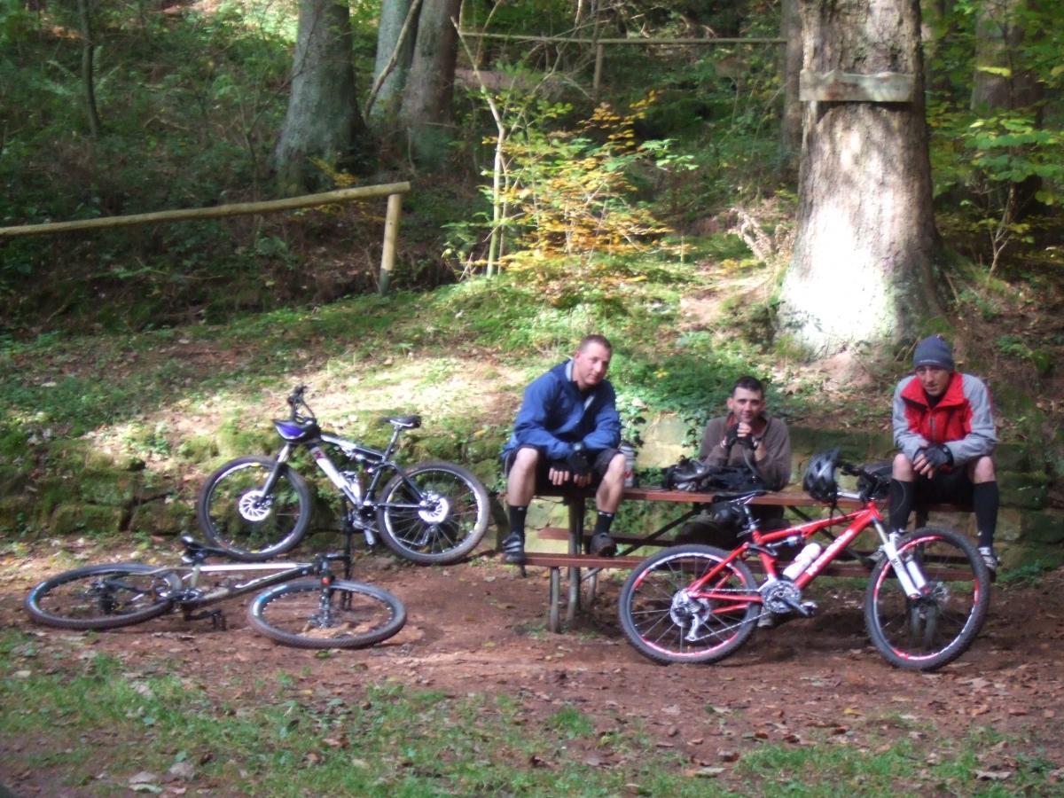 Three men resting on a bench in a forested area, surrounded by trees and greenery. Two mountain bikes are positioned nearby, with one bike lying on the ground. The men are casually dressed, with one wearing a blue jacket, one in a brown shirt, and one in a red jacket and hat. The scene captures a peaceful moment during their biking trip. Rodalben F Trail mountain bike trail.