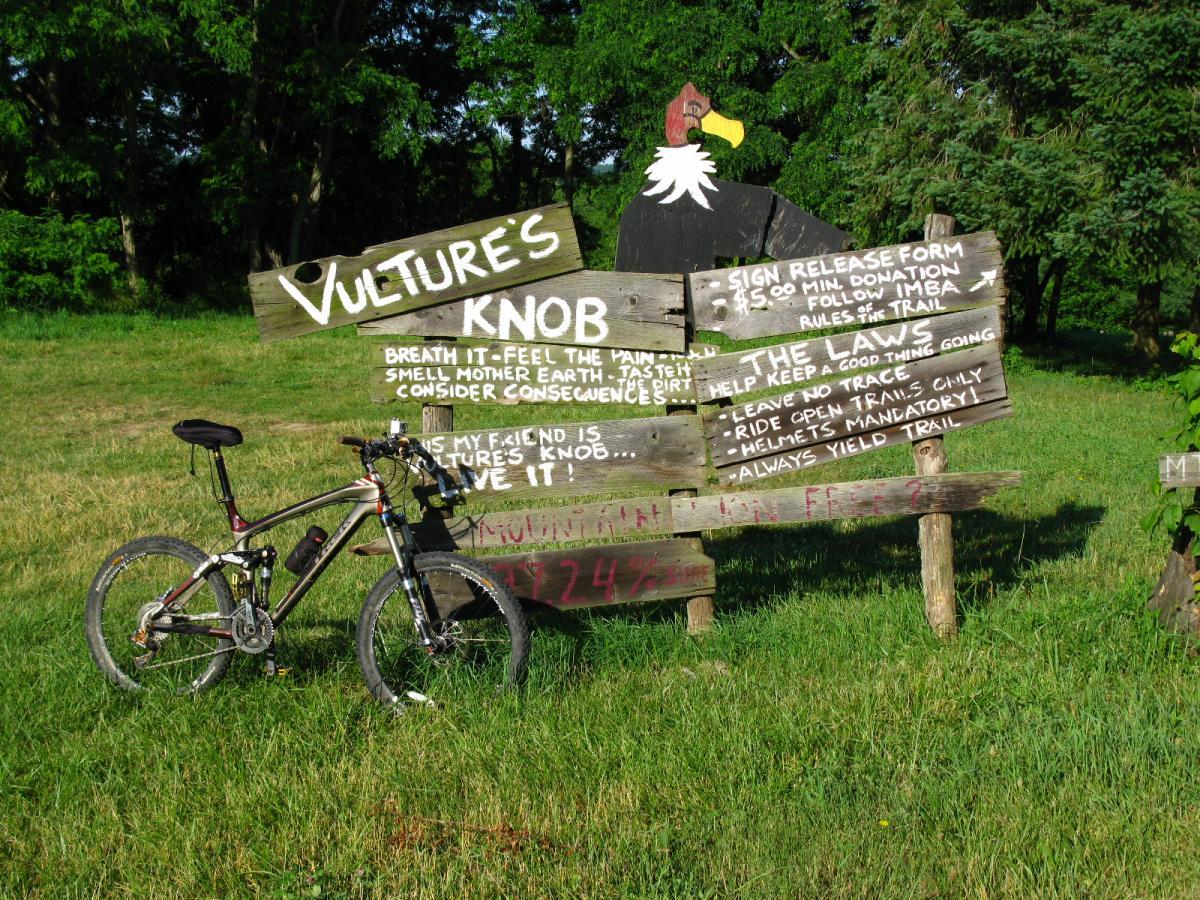 An outdoor sign for "Vulture's Knob" featuring colorful text and a graphic of a vulture. The sign outlines rules for visitors, including safety guidelines and environmental considerations. In front of the sign, there is a mountain bike resting on the grass, set against a backdrop of trees and open fields. Vultures Knob mountain bike trail.