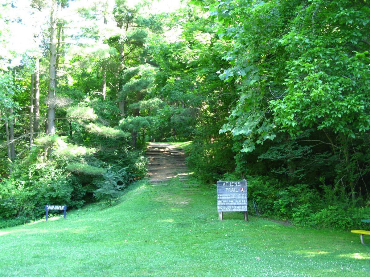 A scenic view of a lush green pathway leading into a wooded area, with a sign on the right indicating "Athens Trail A." The path is bordered by trees and underbrush, suggesting a tranquil outdoor setting for hiking or walking. A second sign can be seen partially on the left. Sells Park mountain bike trail.