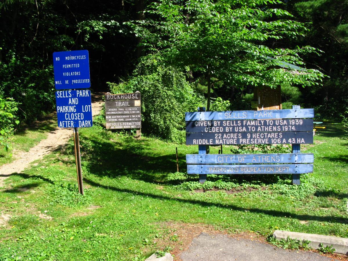 Two signs at the entrance of Sells Park. The blue sign states "No motorcycles permitted; violators will be prosecuted" and notes that the park and parking lot are closed after dark. The brown sign provides information about the Rockhouse Trail and mentions that bicycles are permitted. The area is lush with greenery, indicating a natural outdoor setting. Sells Park mountain bike trail.