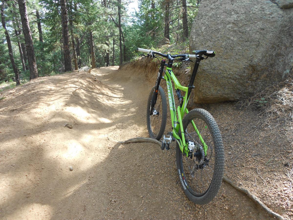 A bright green mountain bike resting on a dirt trail surrounded by trees and boulders. The trail has a slight curve and features a mix of dirt and small rocks, indicating a well-used path through a wooded area. Sunlight filters through the foliage, casting shadows on the ground. Captain Jack's mountain bike trail.