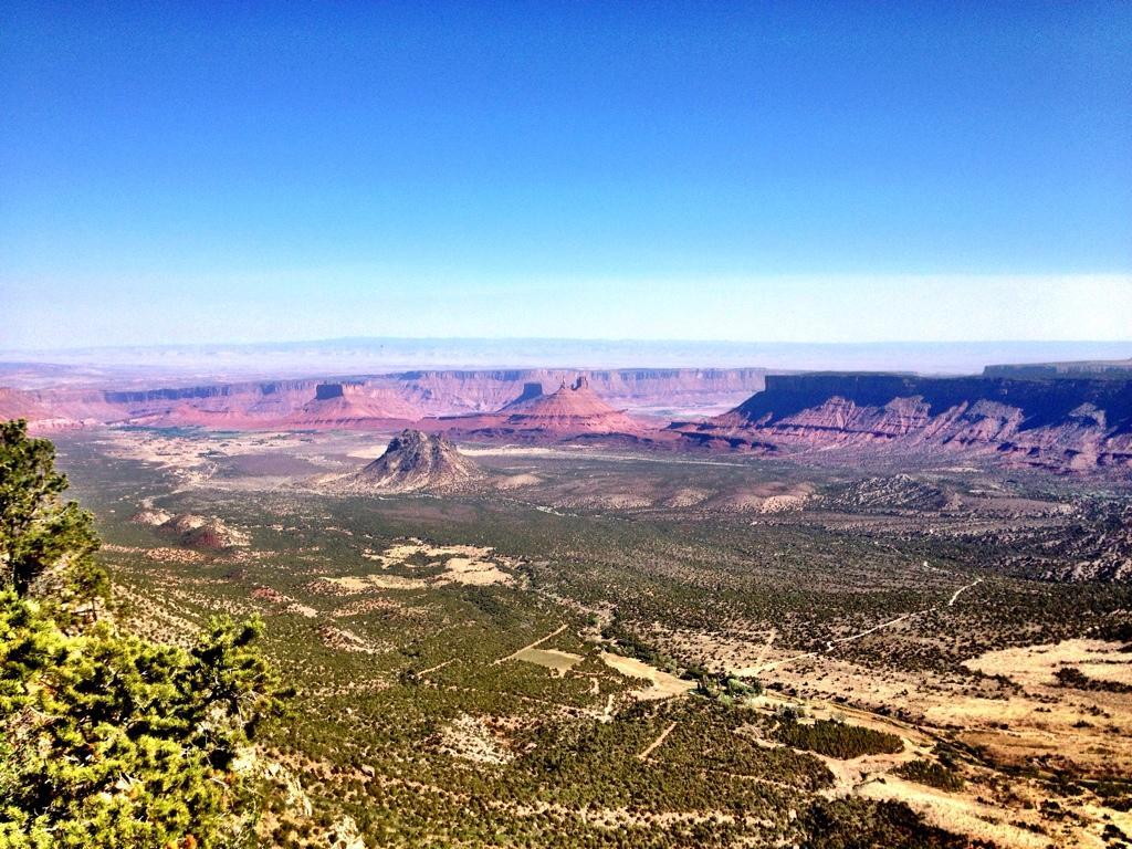 A panoramic view of a vast desert landscape featuring rocky formations and mesas under a clear blue sky. The foreground showcases a mix of greenery and rugged terrain, while distant cliffs and plateaus create a dramatic backdrop. The scene conveys a sense of open space and natural beauty. Porcupine Rim mountain bike trail.