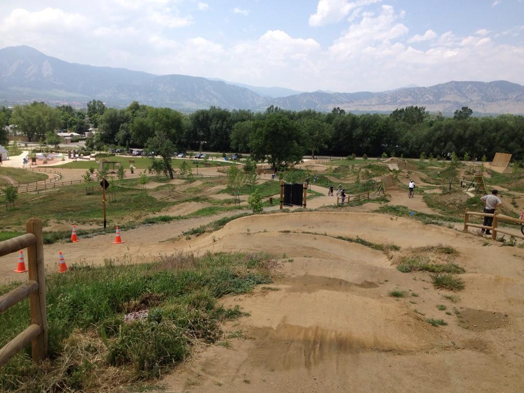 A scenic view of a bike park featuring dirt trails, jumps, and ramps, surrounded by trees and mountains in the background. The area is populated with cyclists riding and maneuvering through various obstacles. Brightly colored traffic cones mark some sections of the park, and the sky is partly cloudy. Valmont Bike Park mountain bike trail.