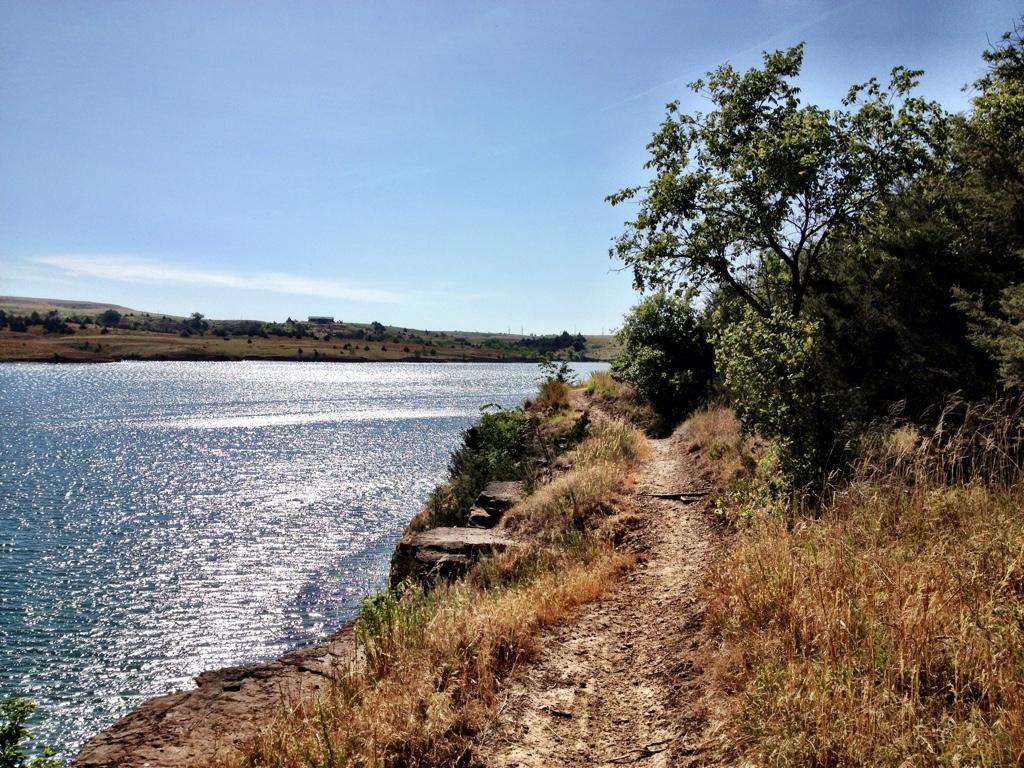 A scenic lakeside path bordered by tall grass and trees, with a calm body of water reflecting sunlight under a blue sky. Switchgrass mountain bike trail.