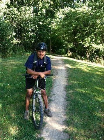 A man wearing a helmet and casual athletic clothing leans on his mountain bike along a dirt path surrounded by greenery. Sunlight filters through the trees in the background, indicating a pleasant outdoor setting. Frick Park mountain bike trail.
