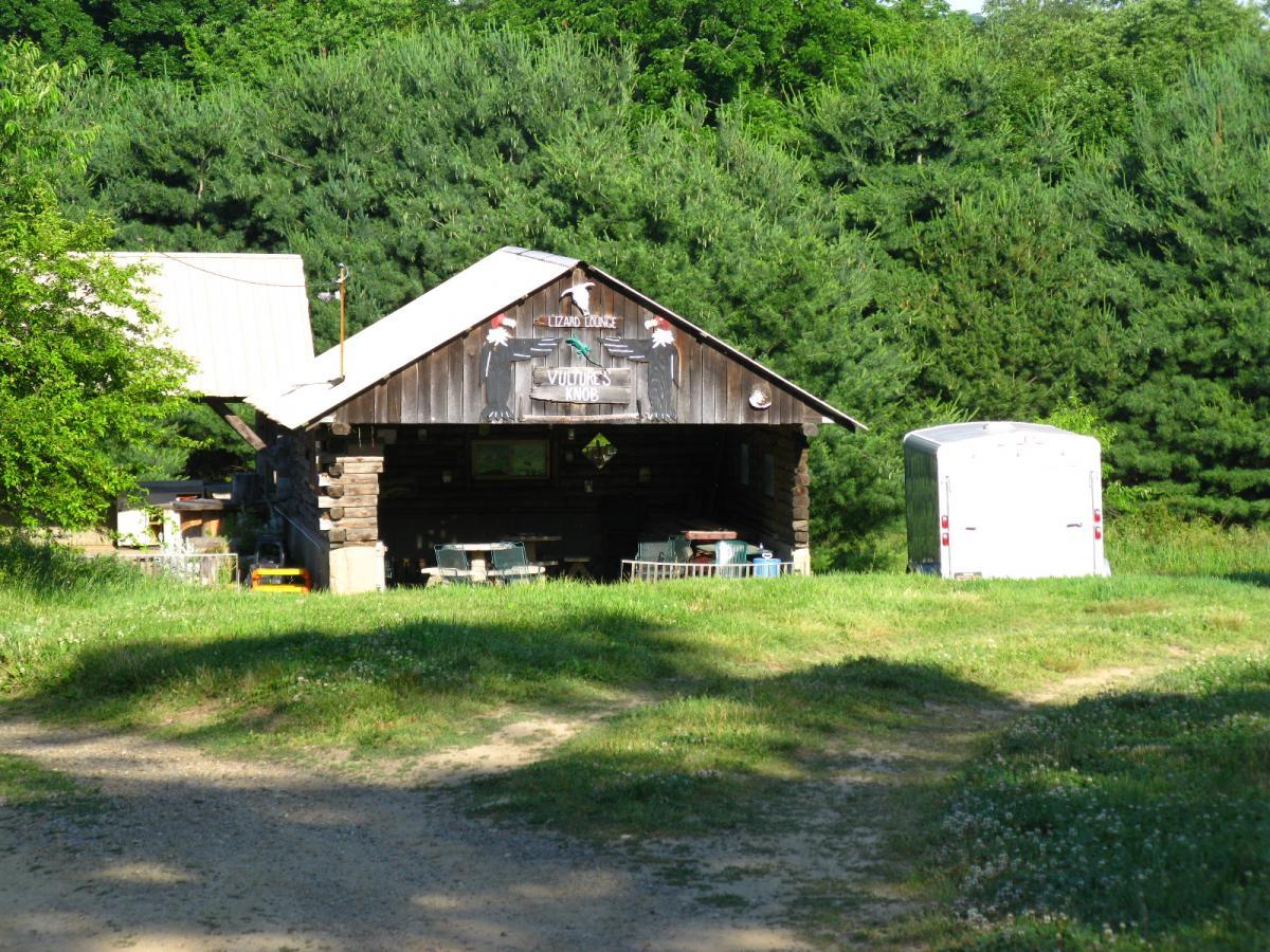 A rustic wooden structure with a sloped roof, labeled "Lizard Lounge," set in a grassy area surrounded by tall trees. A small trailer is parked nearby, and tables and chairs are visible on the porch. The scene is bathed in sunlight, creating a warm and inviting atmosphere. Vultures Knob mountain bike trail.
