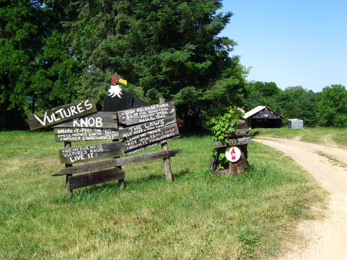 A rustic wooden sign for "Vulture's Knob," surrounded by greenery and leading to a dirt path. The sign includes playful text urging visitors to breathe, feel the earth, and consider the consequences of their actions. There are additional signs with rules and donation information, and a small building is visible in the background. Vultures Knob mountain bike trail.