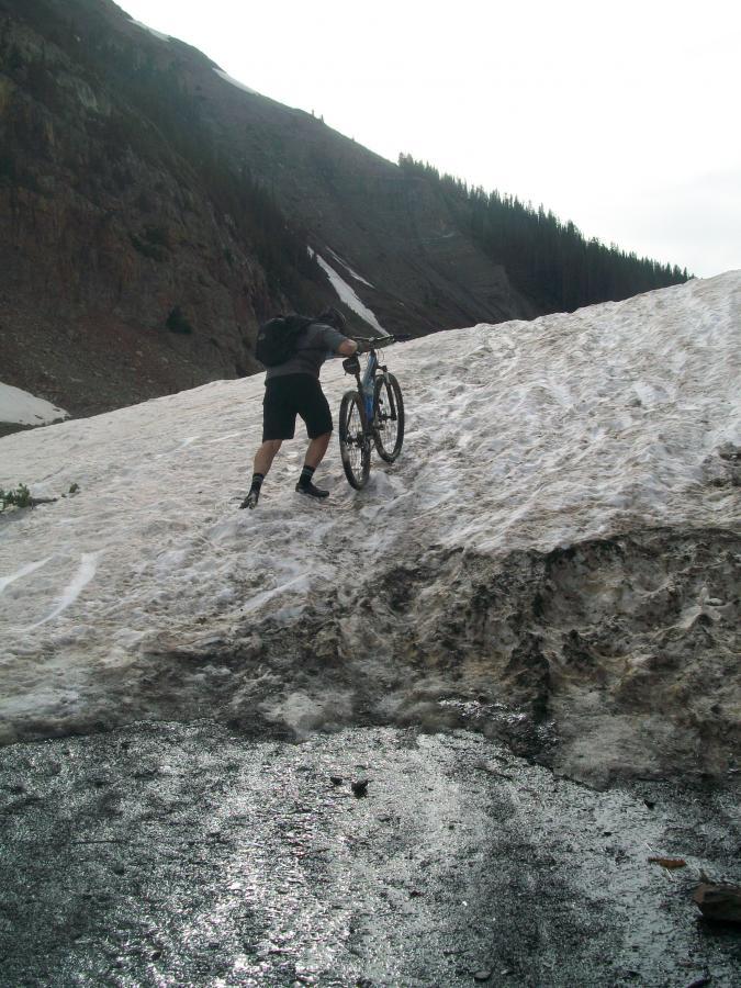 A cyclist pushing a mountain bike uphill over a snowy patch in a mountainous region, with rocky terrain and trees in the background under an overcast sky. Trail 401 mountain bike trail.