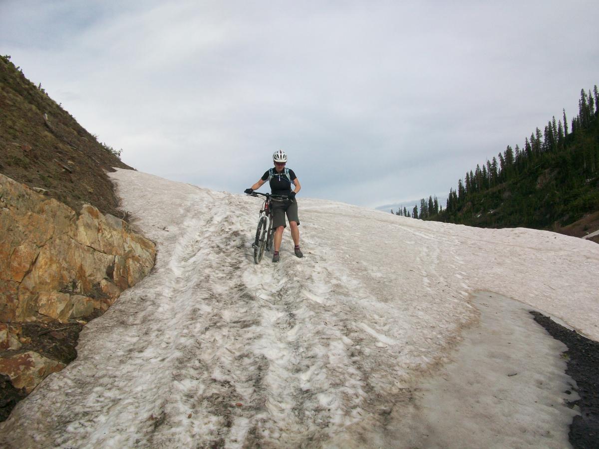 A person riding a mountain bike on a snowy trail in a mountainous area, surrounded by rocky terrain and pine trees under a cloudy sky. Trail 401 mountain bike trail.