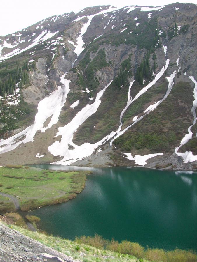 A serene mountain landscape featuring a turquoise lake at the base of rugged slopes. The rocky terrain is interspersed with patches of green vegetation and remnants of snow, highlighting the transition from winter to spring. The scene captures the natural beauty and tranquility of a mountainous region. Trail 401 mountain bike trail.