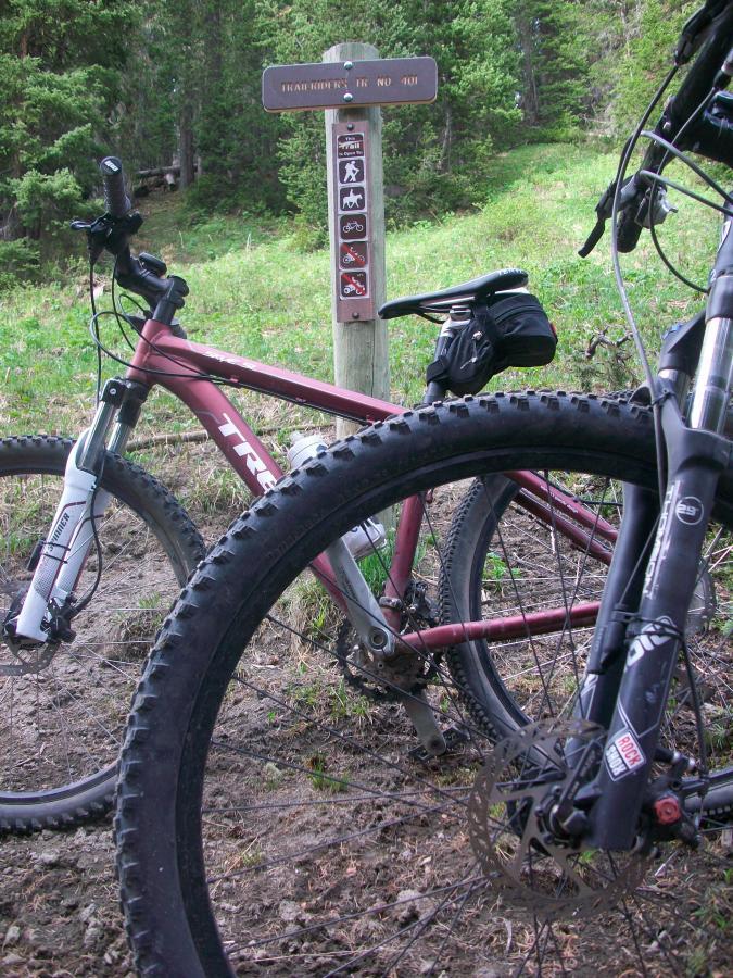 Two mountain bikes are positioned in the foreground, partially obscuring a trailhead sign that provides information about the trail. The sign features symbols indicating various trail uses, including hiking, biking, and horseback riding, amidst a lush green landscape. The background is filled with trees, creating a natural setting. Trail 401 mountain bike trail.