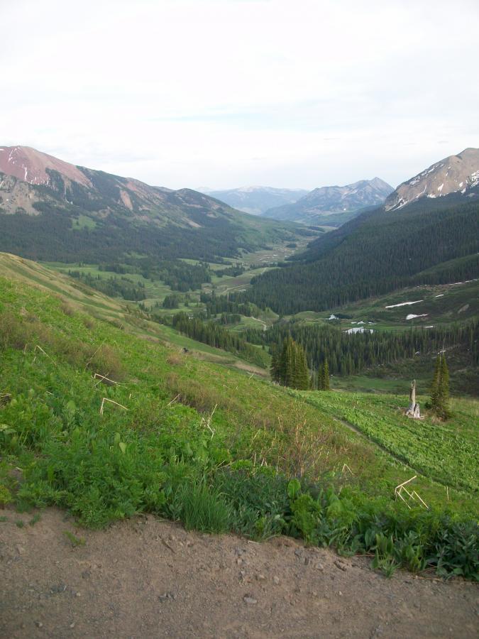 A panoramic view of a lush green valley surrounded by mountains, with patches of snow on the peaks. The foreground features rolling hills covered in grass and small plants, while the backdrop showcases a range of mountains under a cloudy sky. The serene landscape includes a winding river and vibrant tree lines in the valley below. Trail 401 mountain bike trail.