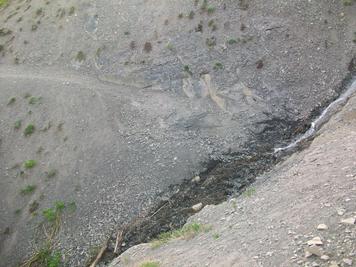 A rocky hillside with patches of green vegetation, showing a narrow trail winding down the slope. At the bottom, a small stream of water flows over a rocky bed, surrounded by loose stones and earth. The terrain appears rugged and uneven, characteristic of a mountainous area. Trail 401 mountain bike trail.