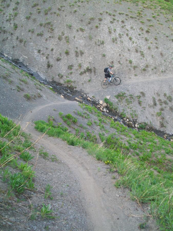 A mountain biker navigates a winding trail along a hillside, with a small creek visible below. The landscape features patches of green grass and rocky terrain. Trail 401 mountain bike trail.