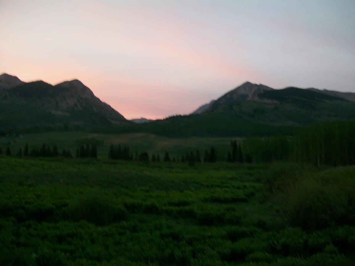 A tranquil landscape featuring mountains under a pastel sky at dusk, with rolling green hills and patches of trees in the foreground. Trail 401 mountain bike trail.