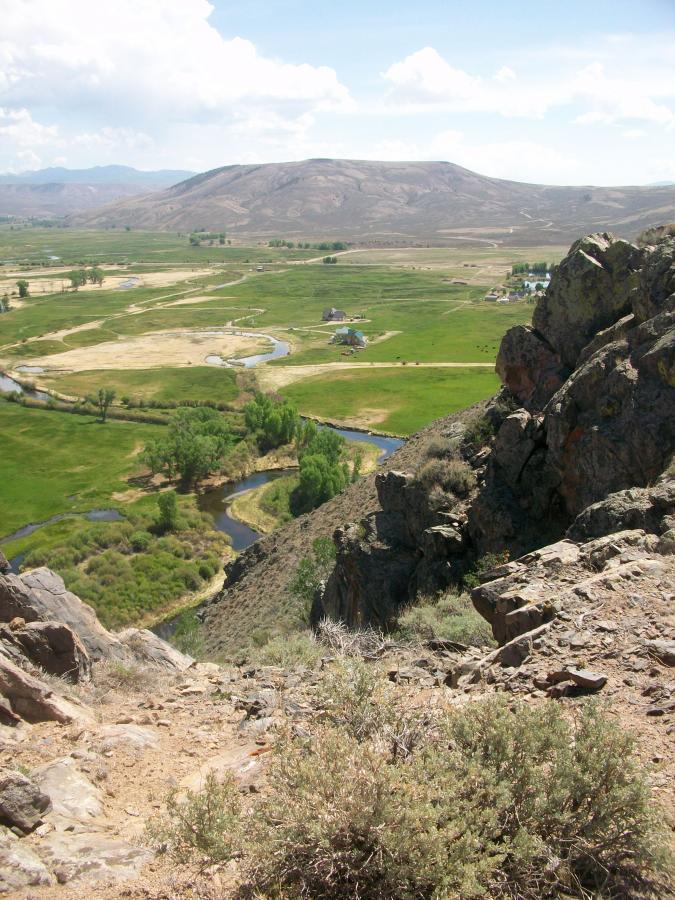 A panoramic view from a rocky cliff overlooking a lush green valley. The landscape features a winding river, fields, and scattered homes, with distant mountains under a partly cloudy sky. Shrubs and rocky terrain are visible in the foreground, highlighting the natural settings. Hartman Rocks mountain bike trail.