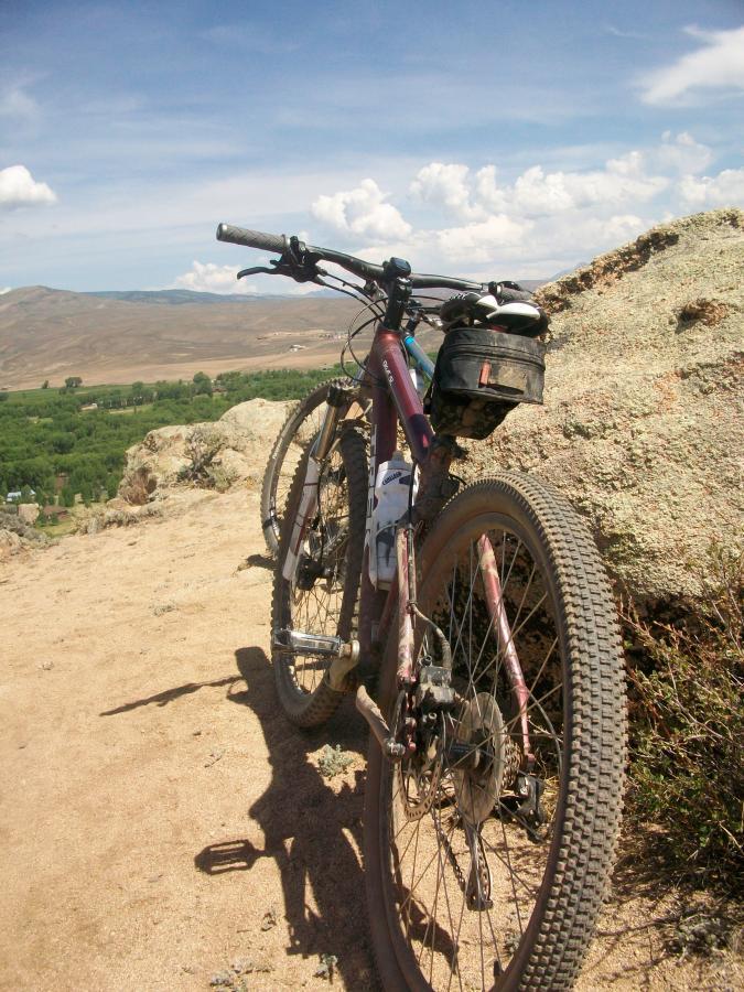 A mountain bike leaning against a rock on a sandy terrain, with a vast landscape of hills and greenery in the background under a partly cloudy sky. Hartman Rocks mountain bike trail.