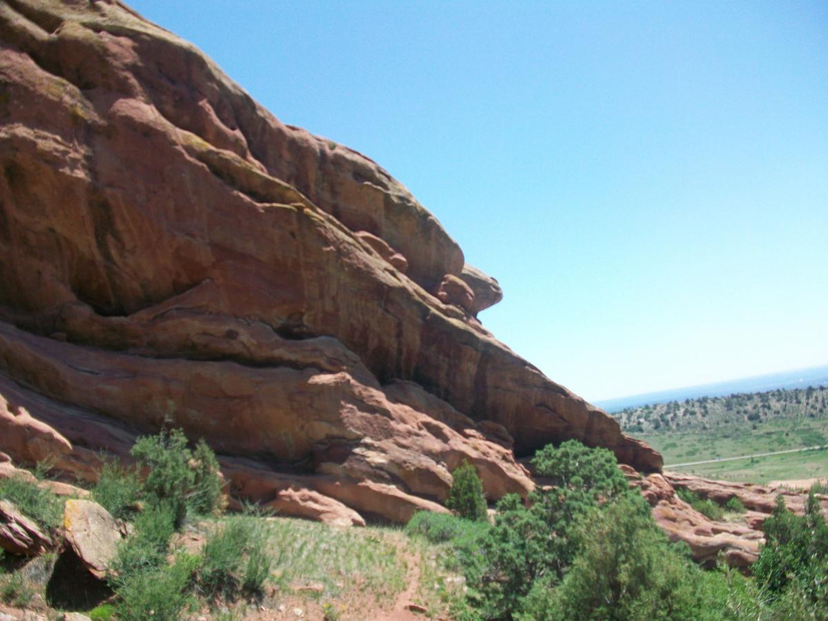 A large red rock formation rises against a clear blue sky, surrounded by green vegetation and rolling hills in the background. The rock displays layered textures, showcasing natural erosion patterns. Red Rocks / Dakota Ridge mountain bike trail.