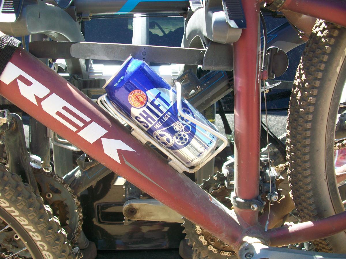 A close-up image of a mountain bike with a blue can of Shift pale lager secured in a bottle holder on the frame. The bike features a maroon color with visible branding, chain, and tires in the background. Sunlight highlights the details of the bike and the can. Buffalo Creek mountain bike trail.