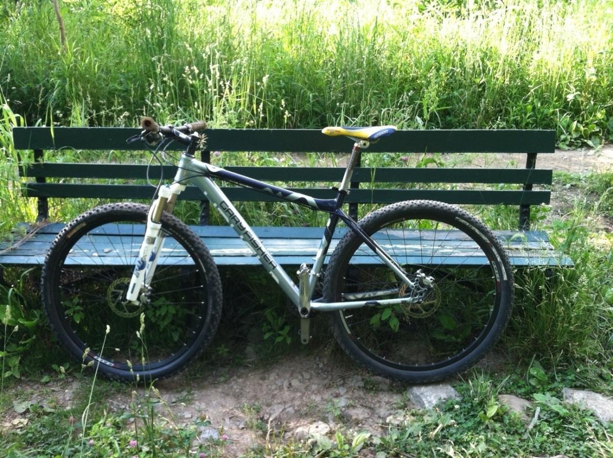 A mountain bike parked beside a green wooden bench, surrounded by tall grass and plants in a natural outdoor setting. Frick Park mountain bike trail.