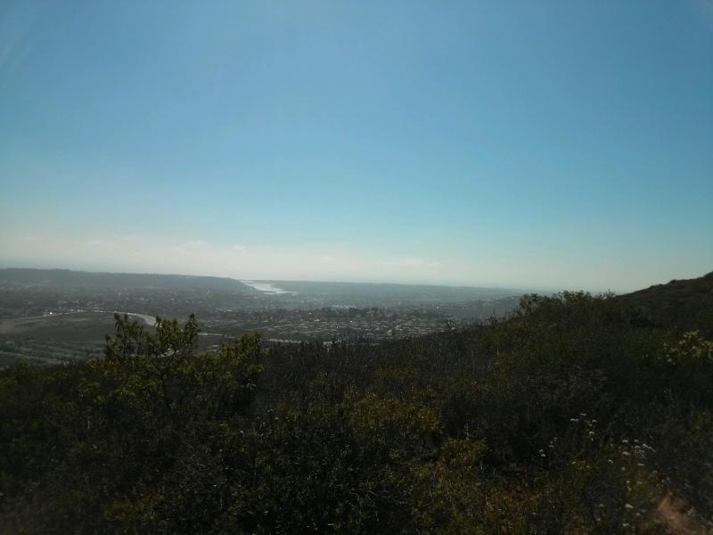 A panoramic view from a hilltop overlooking a cityscape, with green vegetation in the foreground and a clear blue sky above. The horizon features a river winding through the landscape. Rancho La Costa mountain bike trail.