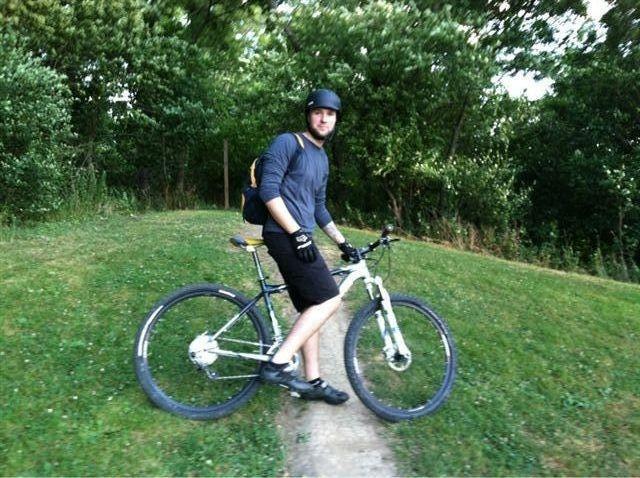 A person wearing a helmet and gloves is sitting on a mountain bike on a grassy path. They are dressed in a long-sleeve shirt and shorts, with a backpack slung over one shoulder. Lush greenery surrounds the area, suggesting a forested or natural setting. Frick Park mountain bike trail.