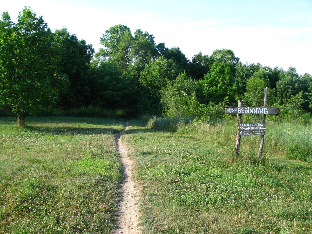 A dirt path leads into a lush, green forest, flanked by tall trees and soft grass. A wooden sign at the path's entrance reads "The Beginning," with additional information below it, suggesting outdoor activities. The scene is bright and inviting, capturing the tranquil beauty of nature. Vultures Knob mountain bike trail.