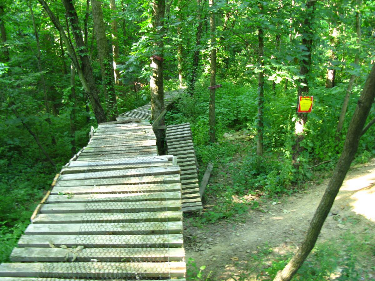 A wooden bike trail winding through a lush green forest, featuring metal mesh for grip on the boards. The path is surrounded by dense foliage, with trees lining either side. A warning sign is visible on a nearby tree, indicating trail information. Vultures Knob mountain bike trail.