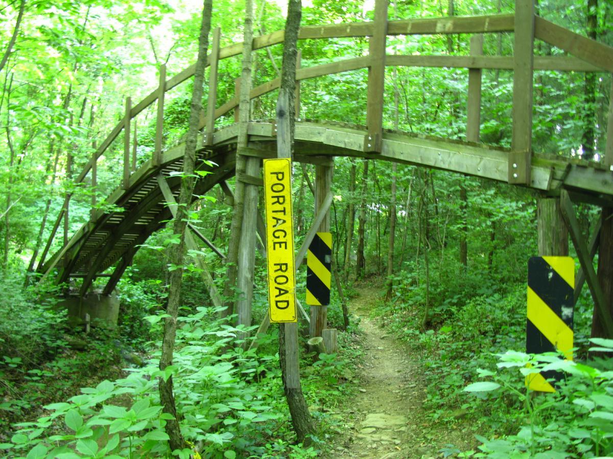 A wooden bridge arches over a lush, green path in a dense forest. A yellow sign reading "PORTAGE ROAD" stands next to the bridge, while black and yellow warning signs are positioned on either side of the entrance to the path. The scene is framed by vibrant greenery, creating a tranquil and natural atmosphere. Vultures Knob mountain bike trail.