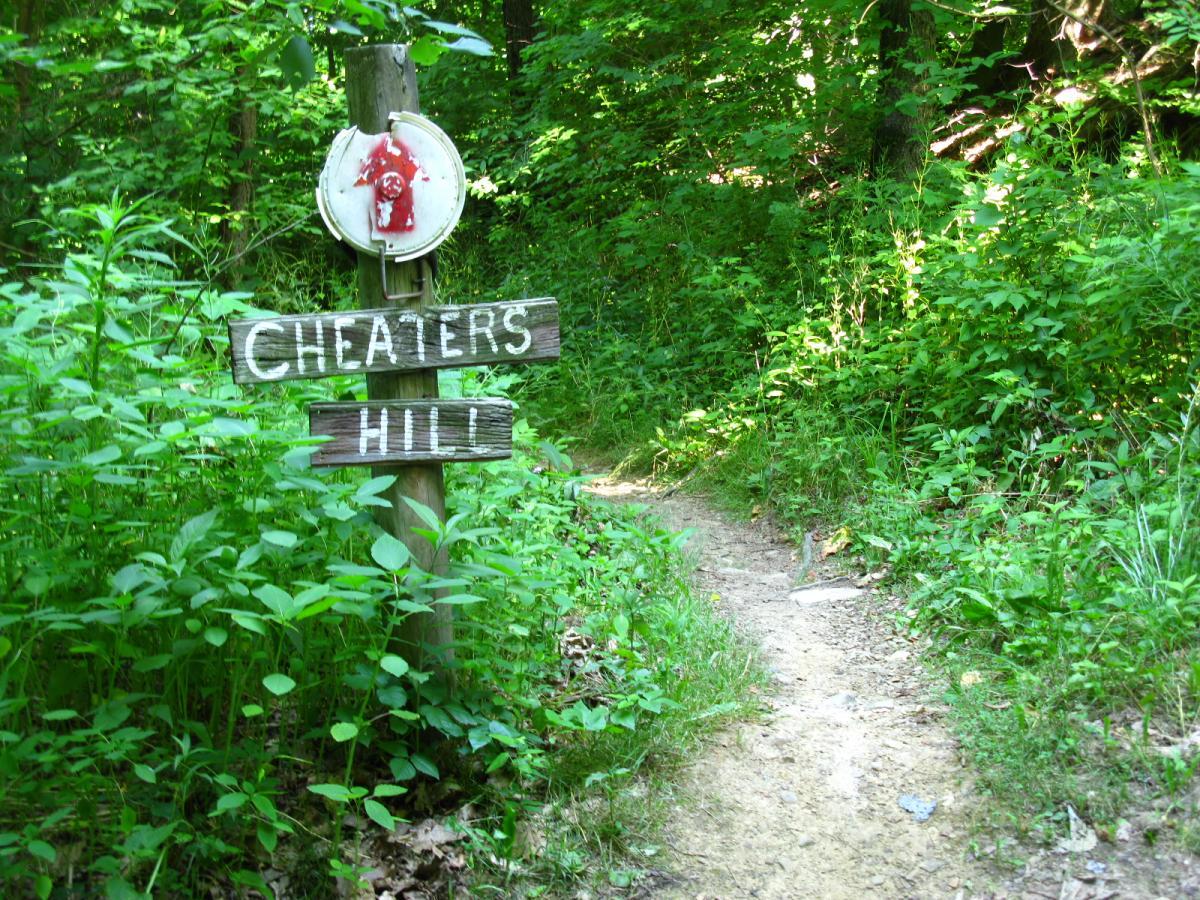 A wooden sign labeled "Cheaters Hill" stands at a trailhead surrounded by lush green foliage. The path, partially visible, leads into a wooded area. The sign has a weathered look, indicating it has been in place for some time. Vultures Knob mountain bike trail.
