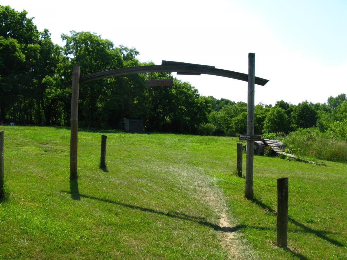 A wooden archway marking the entrance to a grassy path, surrounded by trees. The arch features a sign above it, partially obscured, indicating the direction. The scene is bright and sunny, highlighting the lush greenery of the area. Vultures Knob mountain bike trail.