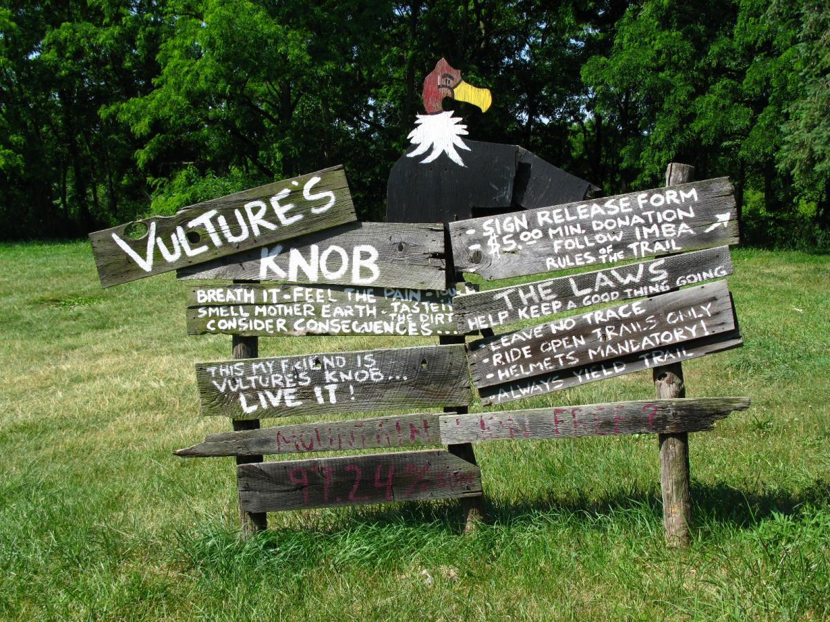 A weathered, wooden sign for Vulture's Knob, featuring various handwritten messages and guidelines. The sign includes phrases encouraging visitors to connect with nature, details about signing a release form for trail use, and rules such as "Leave No Trace" and "Helmets Mandatory." The background features green grass and trees, creating an inviting outdoor atmosphere. Vultures Knob mountain bike trail.