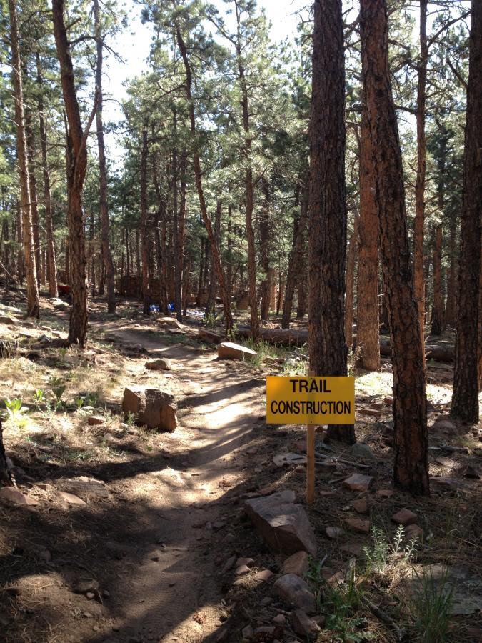 A dirt trail winding through a forest of tall pine trees, with a yellow sign that reads "TRAIL CONSTRUCTION" positioned at the side of the path. The ground is rocky and uneven, and sunlight filters through the tree branches. Heil Valley Ranch mountain bike trail.
