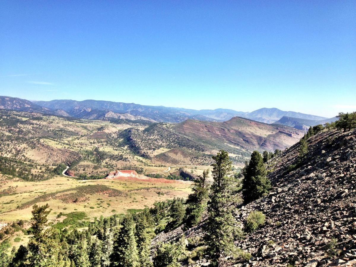 A panoramic view of a mountainous landscape featuring rolling hills, a winding river, and patches of greenery. The sky is clear and blue, with distant mountains visible on the horizon. Rocky terrain is prominent in the foreground, surrounded by coniferous trees. Heil Valley Ranch mountain bike trail.