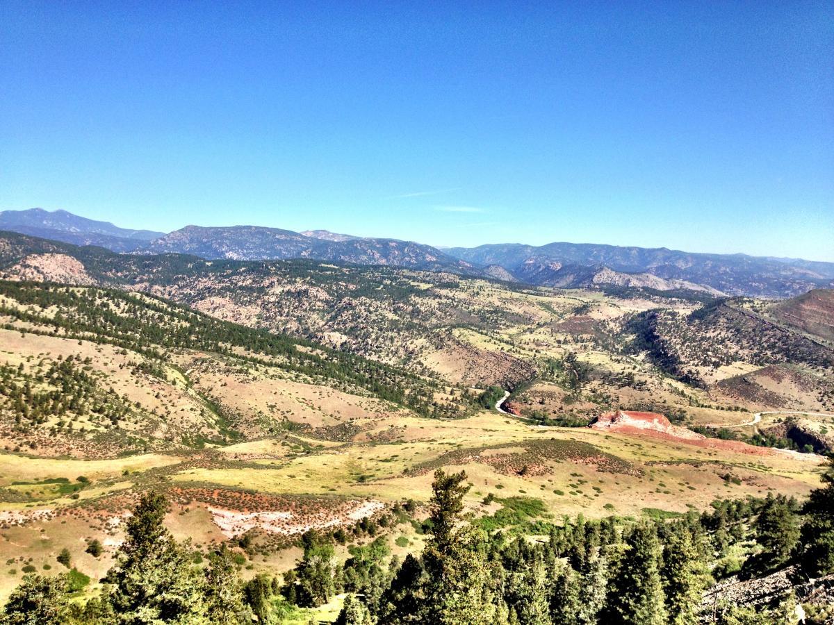 A panoramic view of rolling hills and mountains under a clear blue sky. The landscape features patches of greenery and sparse trees, with a winding river visible in the valley below. The foreground includes various textures of soil and vegetation, while distant mountains provide a scenic backdrop. Heil Valley Ranch mountain bike trail.