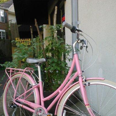 A vintage-style pink bicycle parked against a wall, with some greenery in the background and a sign partially visible. The bike features a rear rack and has a simple, classic design.
